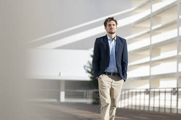 Confident businessman looking away while standing with hands in pockets against office building - SBOF02861