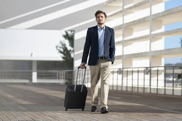 Businessman with luggage looking away while walking on footpath - SBOF02862