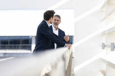 Mature businessman smiling while talking with colleague standing at office building terrace - SBOF02875