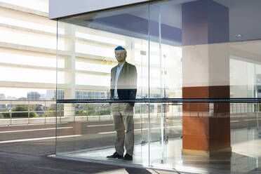 Confident businessman with hands in pockets standing by window at modern office - SBOF02890