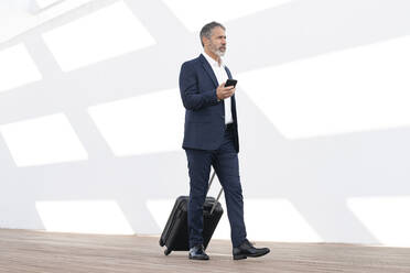 Mature businessman with mobile phone walking with wheeled luggage against white wall - SBOF02901