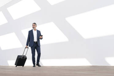 Male entrepreneur holding mobile phone while walking with luggage against white wall - SBOF02903