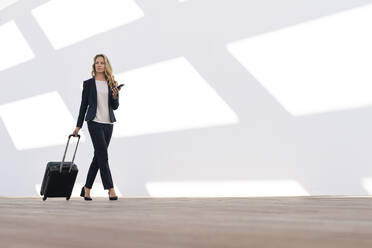 Female entrepreneur with mobile phone and luggage walking against white wall - SBOF02905