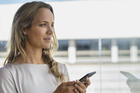 Geschäftsfrau mit Handy, die wegschaut, während sie im Büro steht - SBOF02965