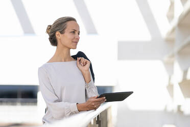 Geschäftsfrau mit digitalem Tablet und Jacke schaut weg, während sie auf einer Büroterrasse steht - SBOF03008