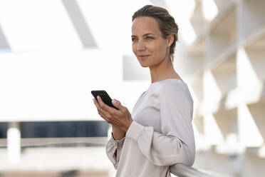 Businesswoman using smart phone while standing at office terrace - SBOF03011