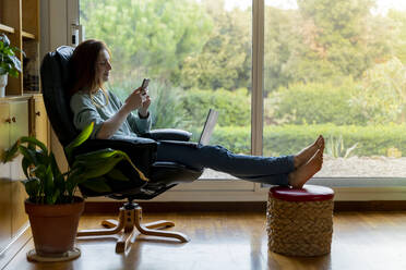 Frau mit Laptop und Mobiltelefon, während sie zu Hause auf einem Stuhl sitzt - AFVF08322