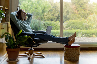 Frau mit Händen hinter dem Kopf entspannt, während sie mit Laptop auf einem Stuhl zu Hause sitzt - AFVF08324