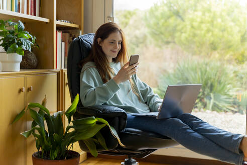 Lächelnde Frau mit Laptop, die ein Smartphone benutzt, während sie zu Hause auf einem Stuhl sitzt - AFVF08325