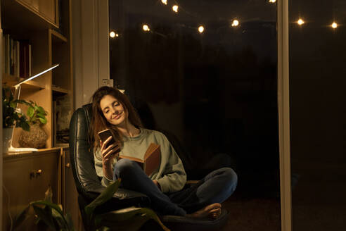Smiling woman with book using smart phone while sitting at home - AFVF08328