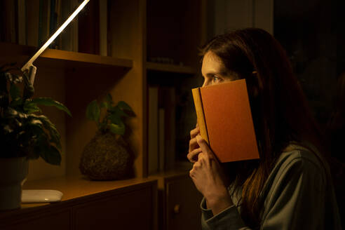 Frau mit Buch starrt auf beleuchtete Lampe, während sie zu Hause sitzt - AFVF08332