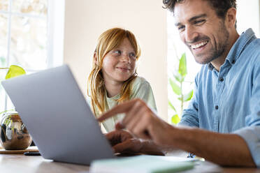 Lächelndes rothaariges Mädchen sieht ihren Vater an, der im Wohnzimmer am Laptop arbeitet - SBOF03012
