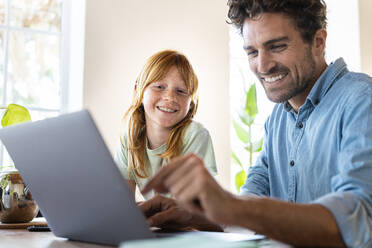 Lächelndes rothaariges Mädchen mit Vater schaut im Wohnzimmer auf einen Laptop - SBOF03013
