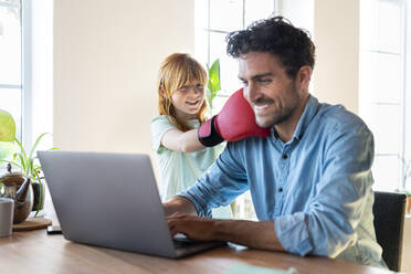 Lächelndes rothaariges Mädchen mit Boxhandschuhen, das seinen Vater schlägt, der zu Hause am Laptop arbeitet - SBOF03021
