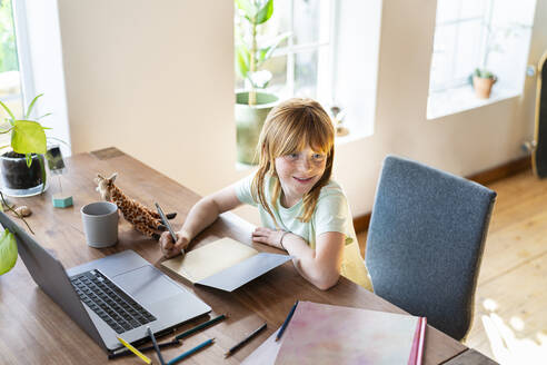 Smiling redhead girl looking away while doing homework in front of laptop at home - SBOF03026