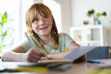 Nettes lächelndes rothaariges Mädchen, das wegschaut, während es zu Hause Hausaufgaben macht - SBOF03027