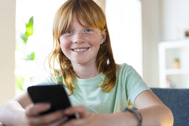 Cute smiling redhead girl holding smart phone at home - SBOF03028