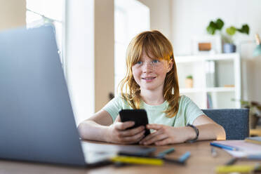 Smiling redhead girl holding smartphone and looking at laptop in living room - SBOF03029