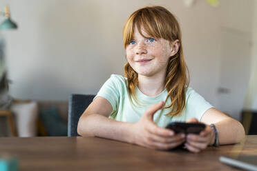 Nettes lächelndes Mädchen mit blauen Augen, das wegschaut, während es ein Smartphone zu Hause hält - SBOF03031