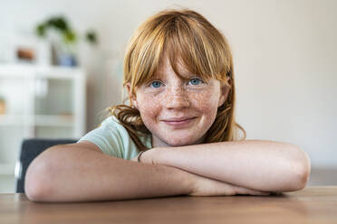 Smiling girl with arms crossed sitting at table in living room - SBOF03032