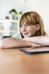 Smiling redhead girl with arms crossed looking away while sitting at table in living room - SBOF03034