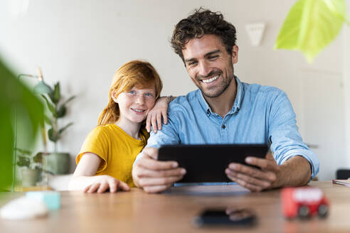 Lächelnder Vater schaut auf ein digitales Tablet, während er neben seiner Tochter zu Hause sitzt - SBOF03037