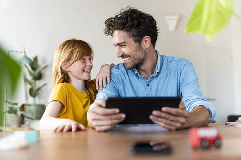Vater mit digitalem Tablet, der seine Tochter anlächelt, während er zu Hause sitzt - SBOF03038