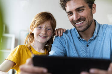 Lächelnder Vater und rothaariges Mädchen schauen auf ein digitales Tablet im Wohnzimmer - SBOF03040