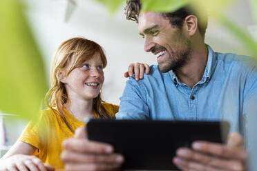 Father and cute redhead daughter smiling at each other while holding digital tablet in living room - SBOF03041