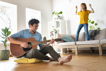 Vater spielt Gitarre und Tochter tanzt auf dem Sofa im Wohnzimmer zu Hause - SBOF03044