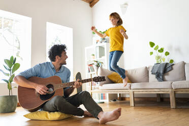 Father playing guitar while daughter jumping from sofa in living room at home - SBOF03045