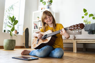 Nettes lächelndes rothaariges Mädchen lernt Gitarre durch digitale Tablet im Wohnzimmer zu Hause - SBOF03047