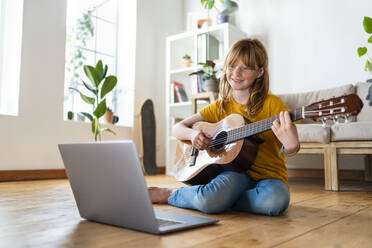 Cute redhead girl learning guitar with online tutorial through laptop in living room at home - SBOF03049