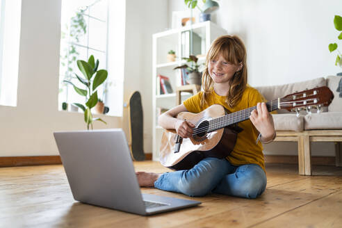 Nettes rothaariges Mädchen lernt Gitarre mit Online-Tutorial durch Laptop im Wohnzimmer zu Hause - SBOF03049