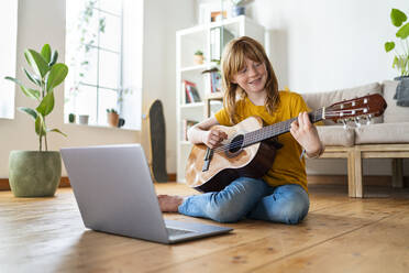 Lächelndes rothaariges Mädchen, das Gitarre spielt, während es zu Hause am Laptop e-Learning betreibt - SBOF03050