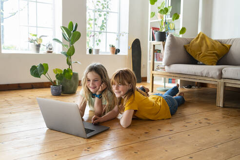 Lächelnde Freundinnen schauen auf einen Laptop, während sie im Wohnzimmer auf der Vorderseite liegen - SBOF03055