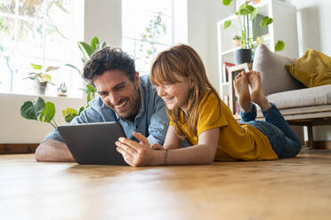 Fröhlicher Vater und Tochter schauen auf ein digitales Tablet, während sie im Wohnzimmer auf dem Boden liegen - SBOF03059