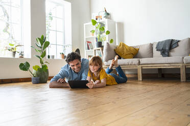 Lächelnder Vater mit Tochter, die auf ein digitales Tablet schaut, während sie zu Hause auf der Vorderseite liegen - SBOF03060