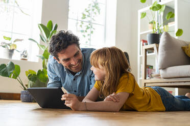 Lächelnde Tochter mit digitalem Tablet, die ihren Vater anschaut, während sie zu Hause im Wohnzimmer auf dem Boden liegt - SBOF03061