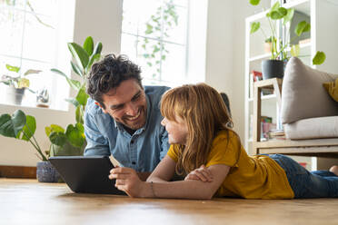 Smiling daughter with digital tablet looking at father while lying on floor in living room at home - SBOF03061