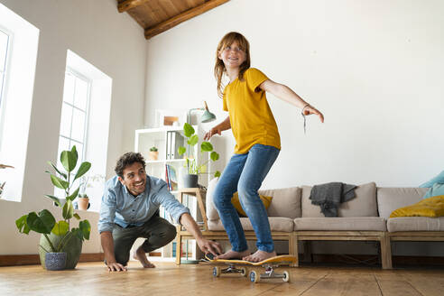 Father helping daughter to ride skateboard in living room at home - SBOF03063