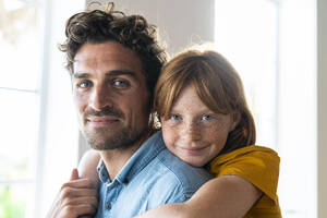 Lächelnde rothaarige Tochter mit blauen Augen kuschelt mit Vater im Wohnzimmer - SBOF03069