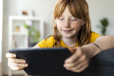 Nettes rothaariges Mädchen schaut auf ein digitales Tablet, während sie auf einem Stuhl im Wohnzimmer sitzt - SBOF03078