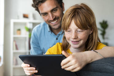 Vater und süße rothaarige Tochter schauen gemeinsam im Wohnzimmer auf ein Tablet - SBOF03079