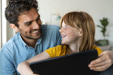 Vater und rothaarige Tochter mit digitalem Tablet lächelnd, während sie sich gegenseitig im Wohnzimmer anschauen - SBOF03080