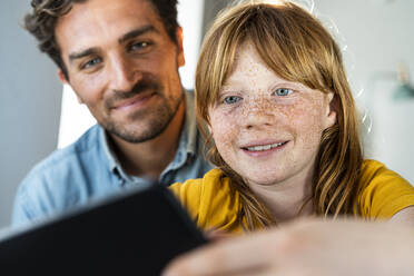 Smiling father and cute redhead daughter looking at digital tablet together in living room - SBOF03081