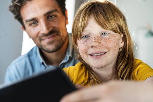 Lächelnder Vater und süße rothaarige Tochter schauen gemeinsam im Wohnzimmer auf ein digitales Tablet - SBOF03081