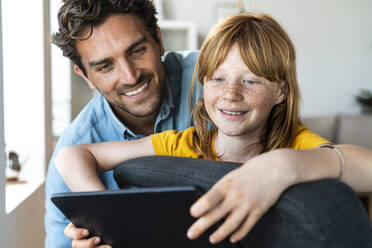 Lächelnder Vater und Tochter schauen gemeinsam auf ein digitales Tablet, während sie auf einem Stuhl zu Hause sitzen - SBOF03082