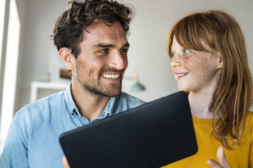 Lächelnde Tochter sieht ihren Vater an, während sie zu Hause ein digitales Tablet hält - SBOF03084