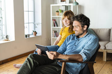 Vater und Tochter schauen auf ein digitales Tablet, während sie zu Hause auf einem Sessel sitzen - SBOF03085